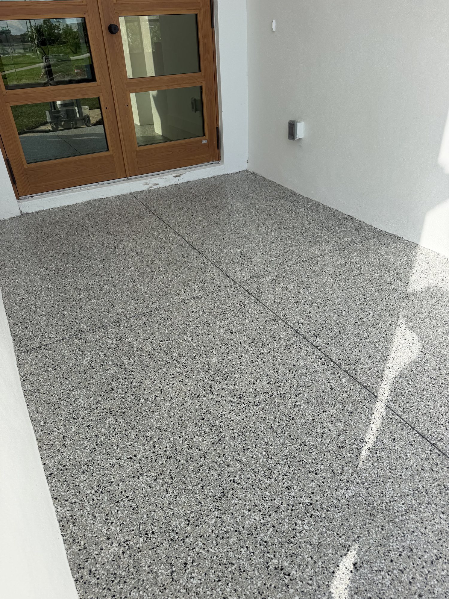 Gray flake coating on an outdoor entry slab with saw-cut joints, running up to a pair of wood-framed glass doors