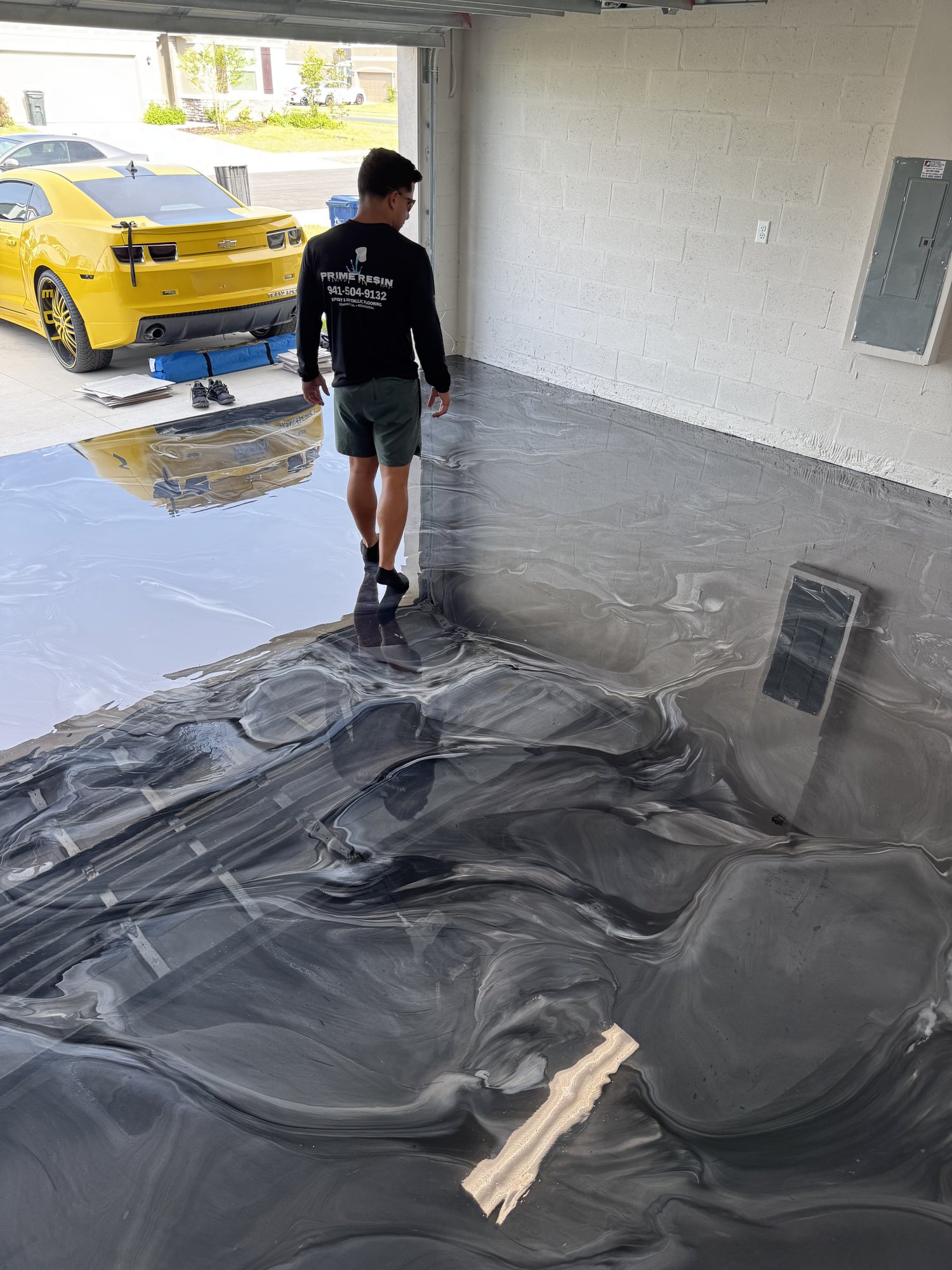 Prime Resin installer in a branded company shirt working a wet charcoal metallic pour, a yellow Camaro on the driveway outside