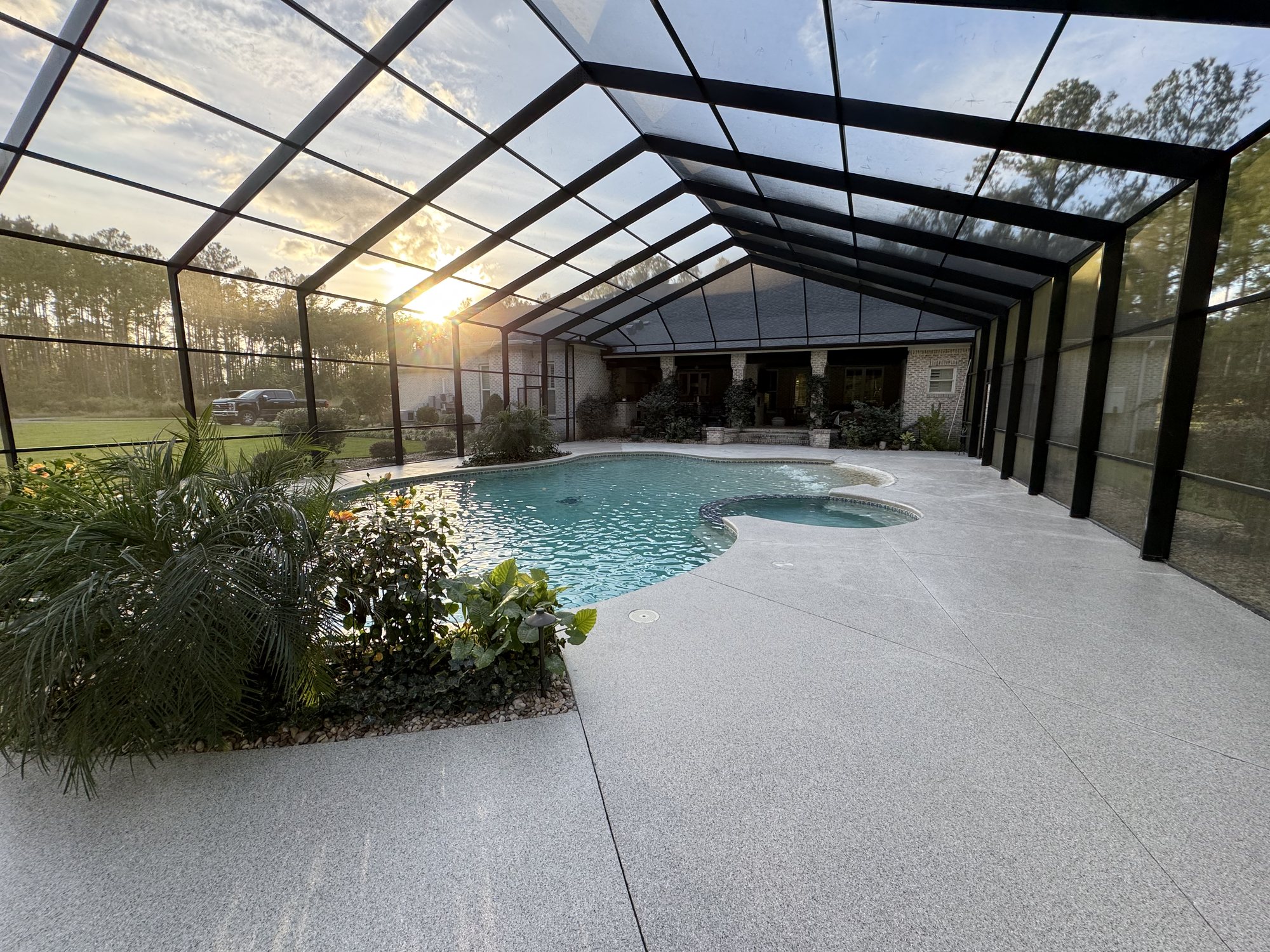 Speckled gray flake pool deck filling a large screened enclosure, sun setting through the screen behind a single-story brick house