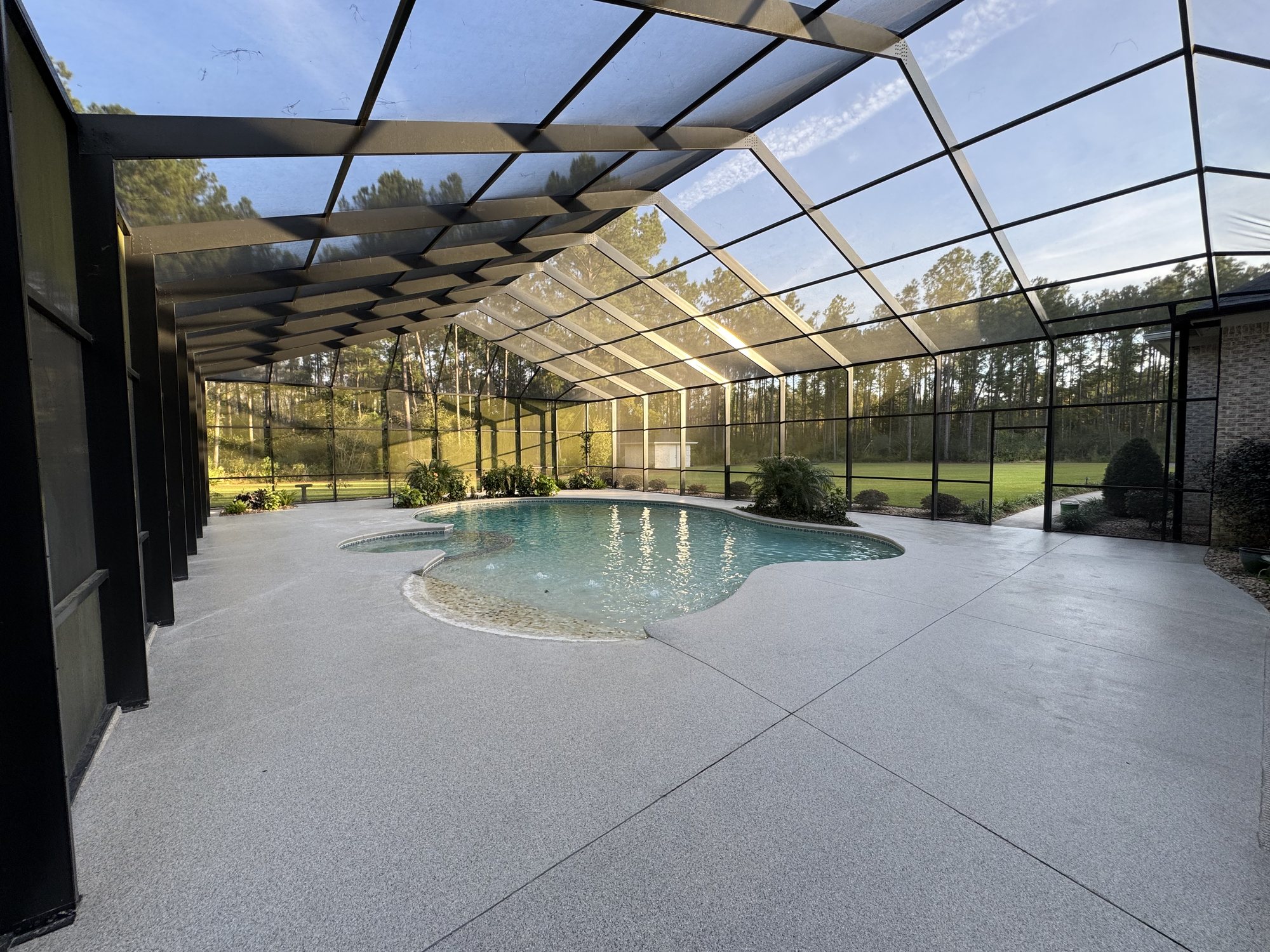Wide flake-coated pool deck under a screened cage, kidney-shaped pool and pines beyond the screen